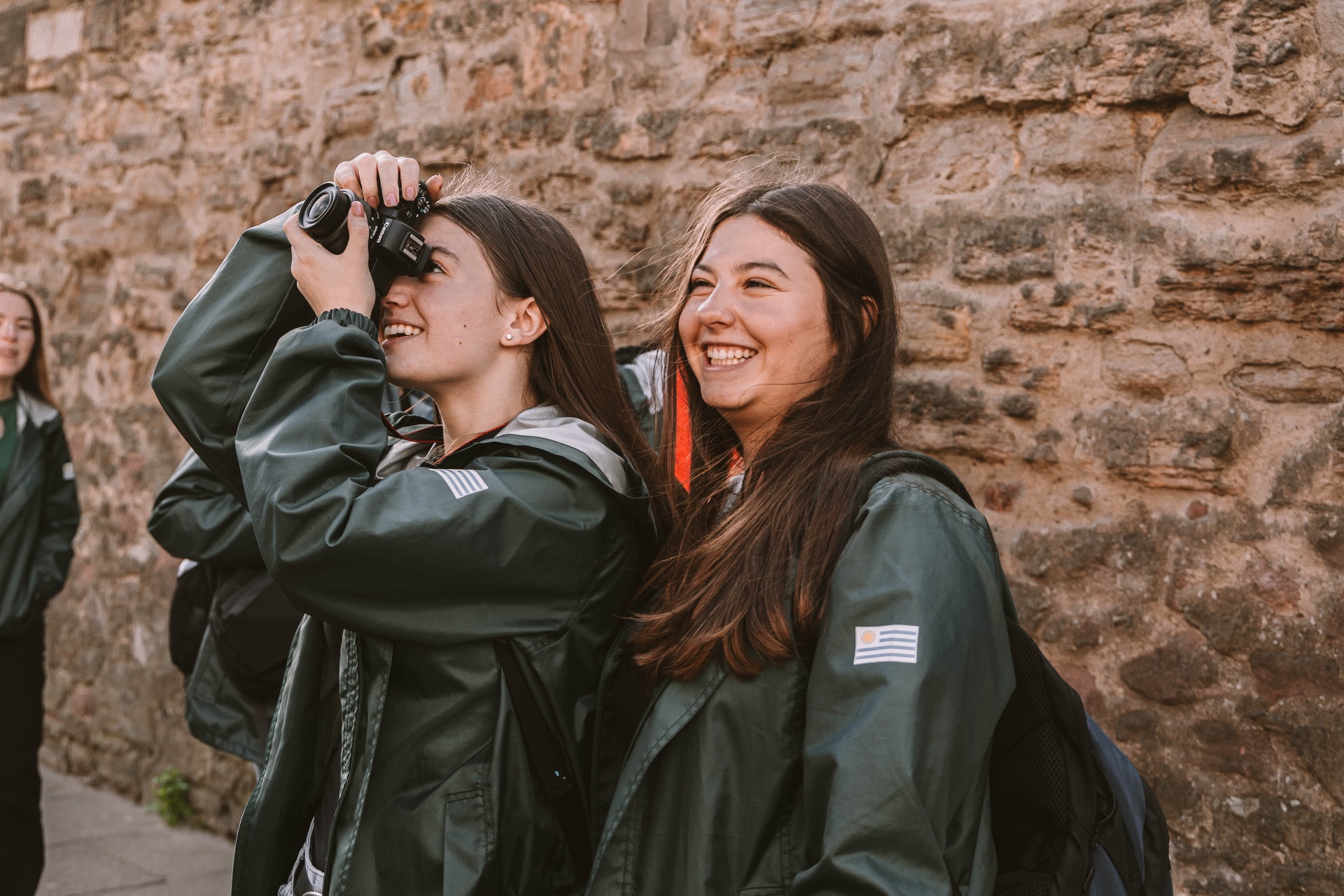 Dos viajeras riendo mientras sacan fotos junto a un muro de piedra
